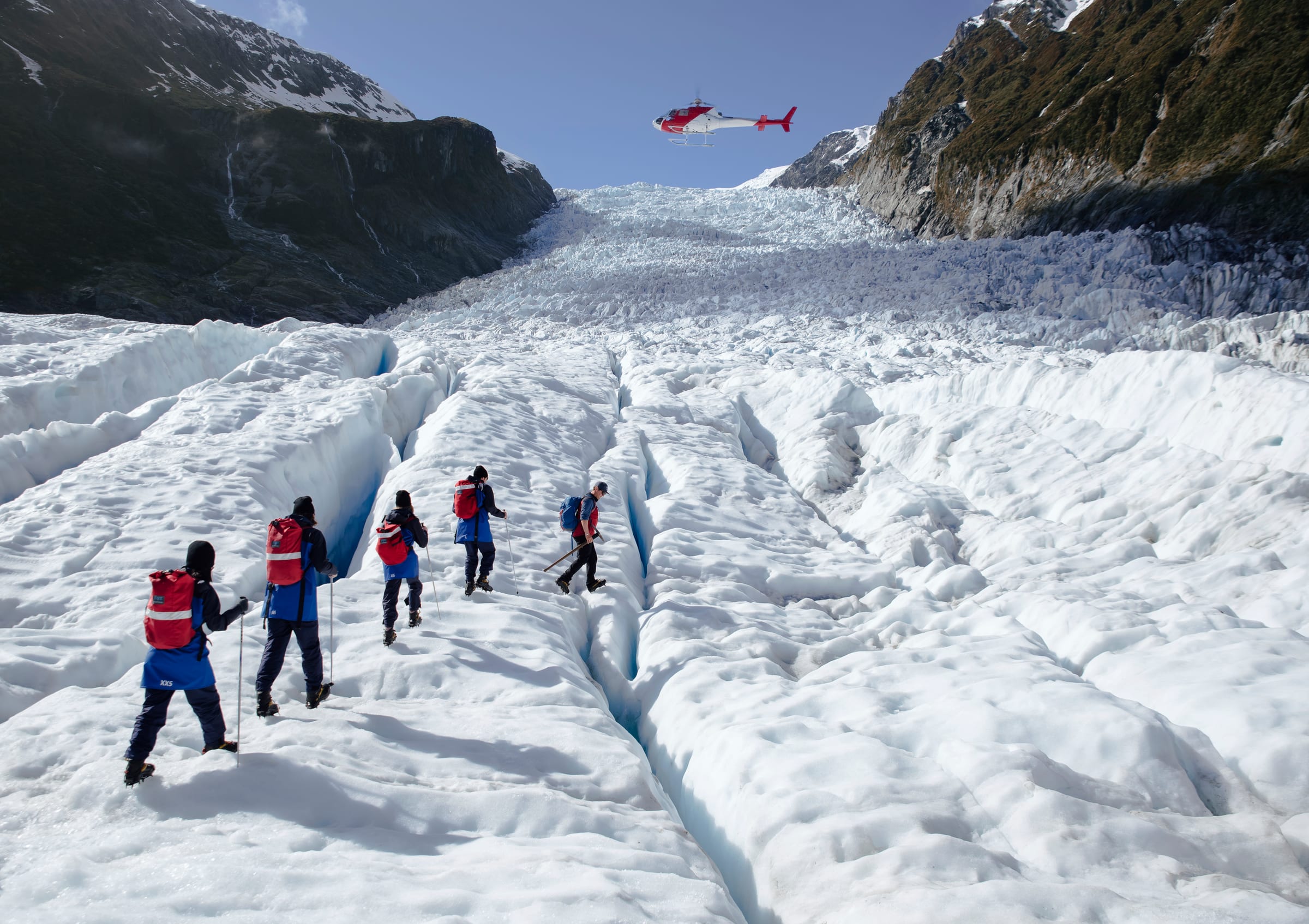 Guided hikers crossing Franz Josef Glacier beneath a helicopter